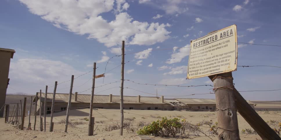 A 'Restricted Area' sign on a barbed wire fence guards an abandoned building in the sandy desert of Kolmanskop, Namibia