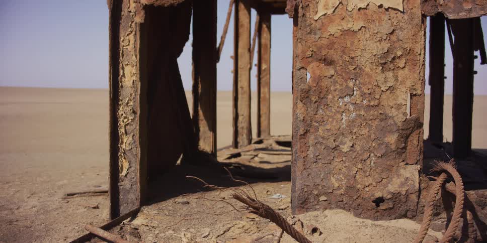 A close-up static shot of a heavily rusted and corroded metal structure abandoned in the vast, sandy desert