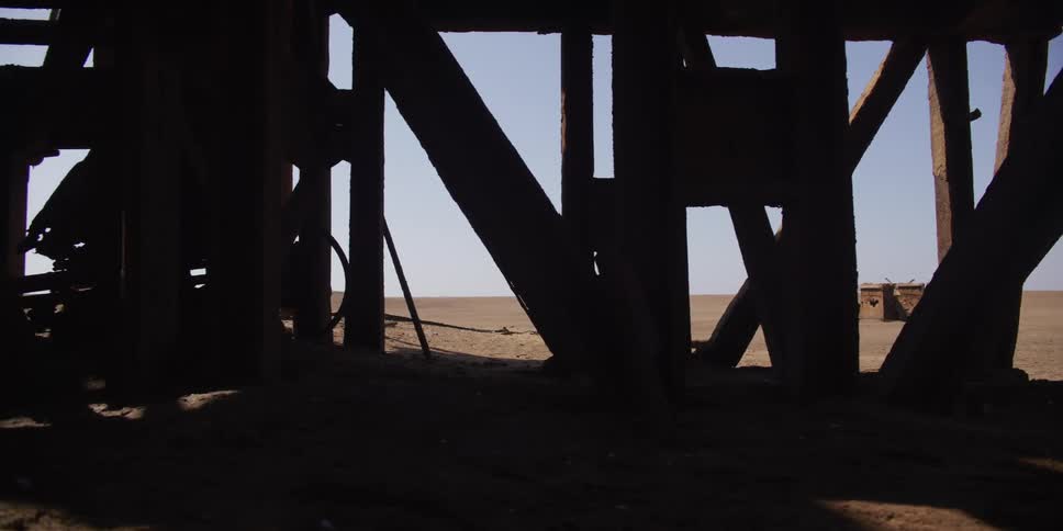 Tracking shot from inside the dark, silhouetted remains of an old wooden trestle looking out at the bright desert