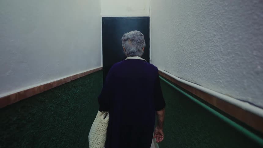 From behind, an elderly woman with a tote bag slowly walks down a narrow, shaded alley, opening a door at the end to step out into a sunlit street