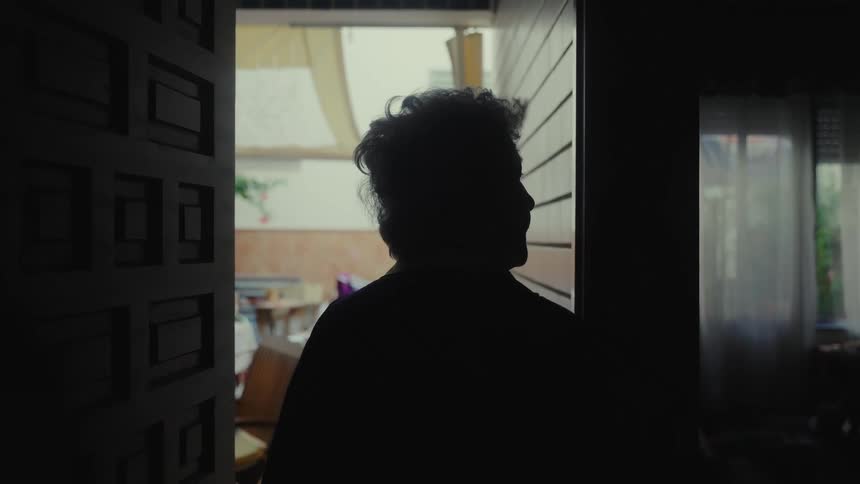 A silhouetted elderly woman slowly walks out of a dark room through an open door into a brighter patio