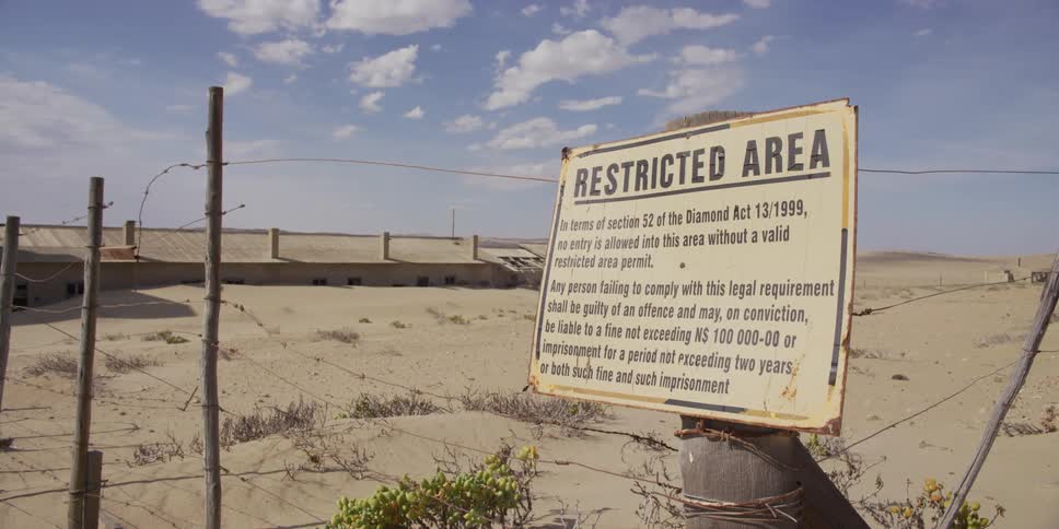 A slow push-in shot of a 'Restricted Area' sign for a diamond mine on a barbed wire fence in a desolate desert with abandoned buildings in the background