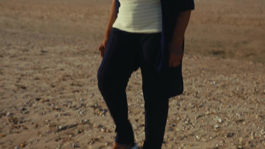 A thoughtful elderly woman enjoys a peaceful walk along a sandy beach on a sunny day in slow motion
