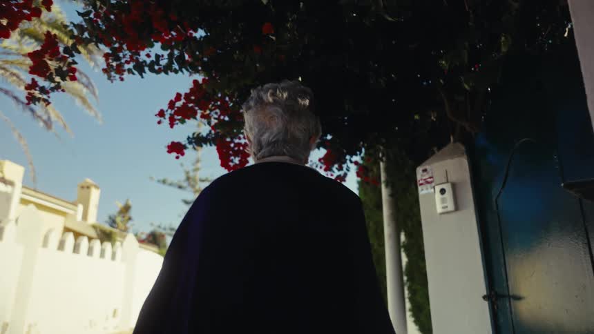 A tracking slow-motion shot follows an elderly woman from behind as she walks down a quiet, sun-drenched street