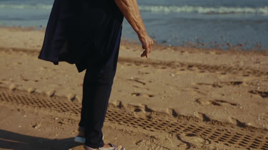 A thoughtful senior woman with grey hair walks along a tranquil beach, gazing at the ocean waves in slow motion