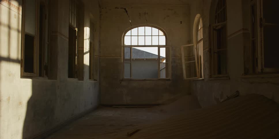 Warm sunlight streams into a decaying room of an abandoned building as a sand dune slowly fills the space