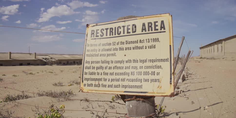 A rusty 'Restricted Area' sign and barbed-wire fence mark the boundary of an abandoned diamond mining town in the desert