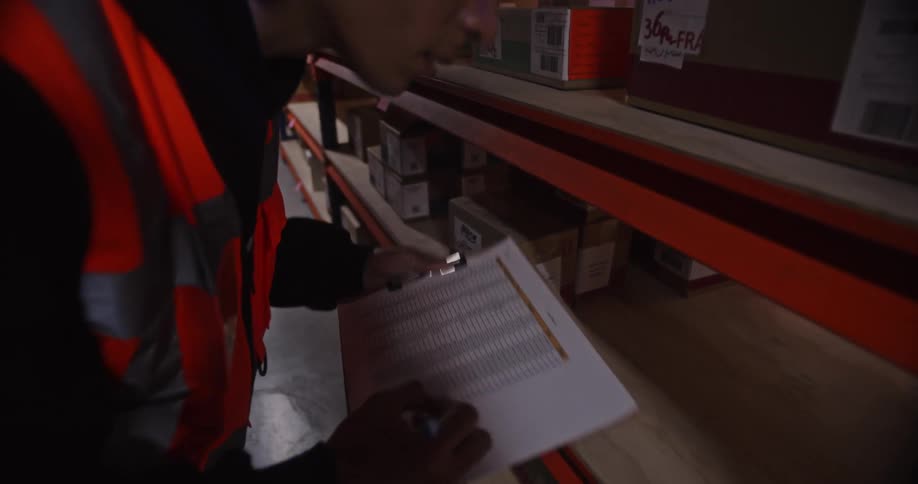 A young warehouse worker walks down an aisle and takes inventory of boxes on a shelf using a clipboard