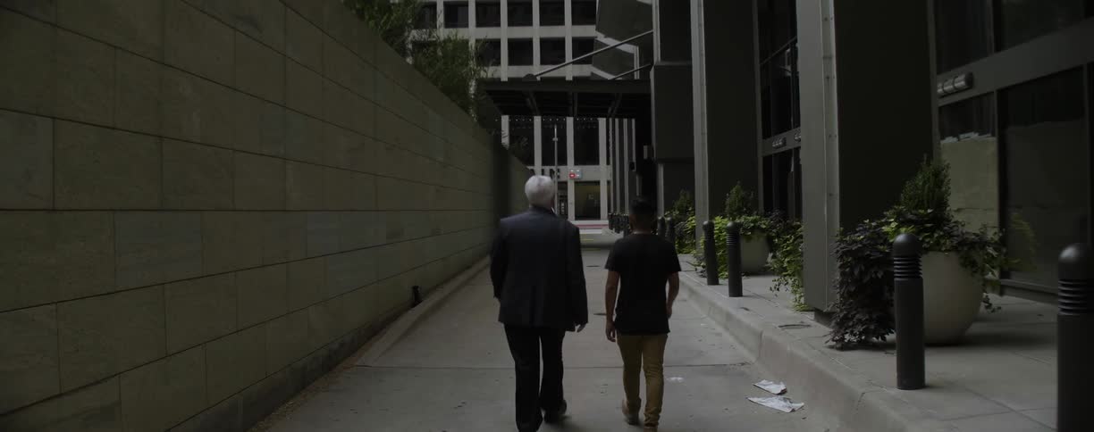 An elderly man and a casually dressed young man walk in between skyscrapers while talking
