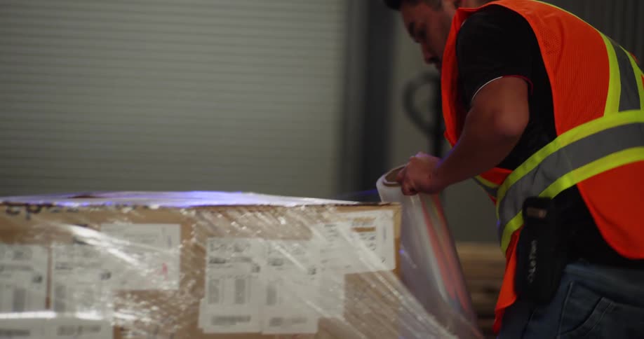 A warehouse worker in a high-visibility vest carefully wraps a pallet of cardboard boxes with plastic shrink wrap in slow motion