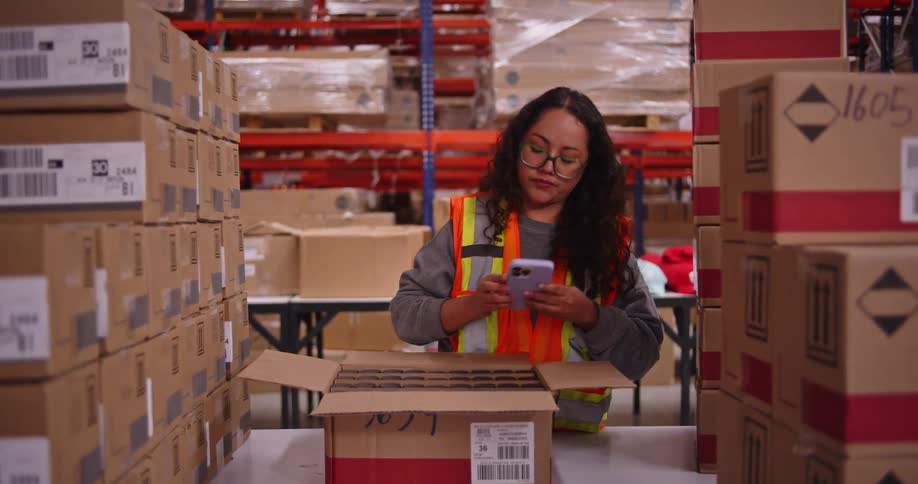 A female warehouse worker uses her smartphone to scan and verify inventory in a cardboard box before closing it