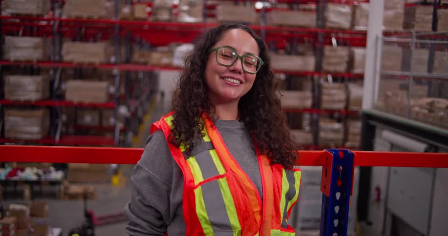 A female warehouse employee in a safety vest smiles confidently for the camera in a large distribution center
