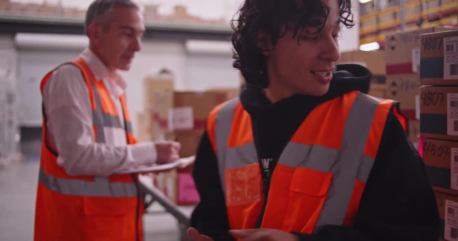 A warehouse supervisor mentors a young employee as they work together checking inventory with a scanner and clipboard in a large distribution center