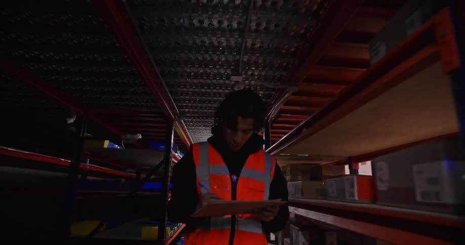 Slow-motion tracking shot of a young warehouse worker in a safety vest taking inventory as he walks down an industrial aisle