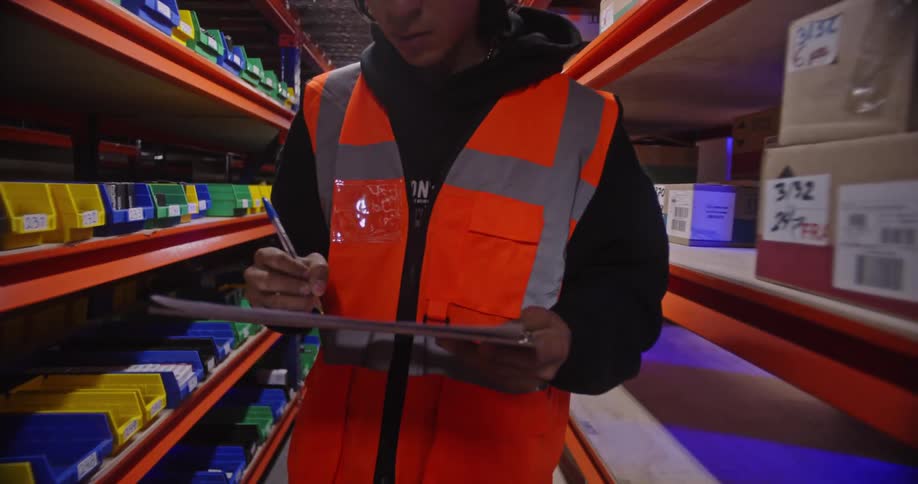 A focused young warehouse employee conducts an inventory check with a clipboard before walking down a long, well-stocked aisle