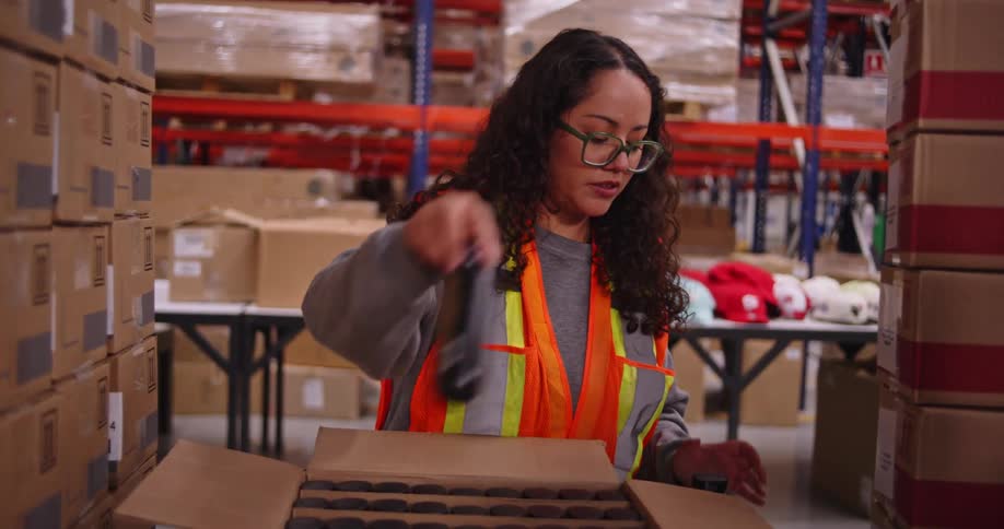 A female warehouse worker meticulously counts and inspects small bottles from a cardboard box in a large distribution center