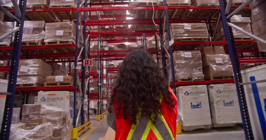 A warehouse worker walks down an aisle lined with towering shelves packed with pallets of goods in a large distribution center