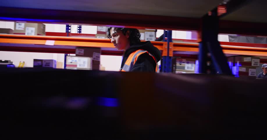 Tracking shot follows a young male employee in a safety vest walking down an aisle in a dimly lit warehouse