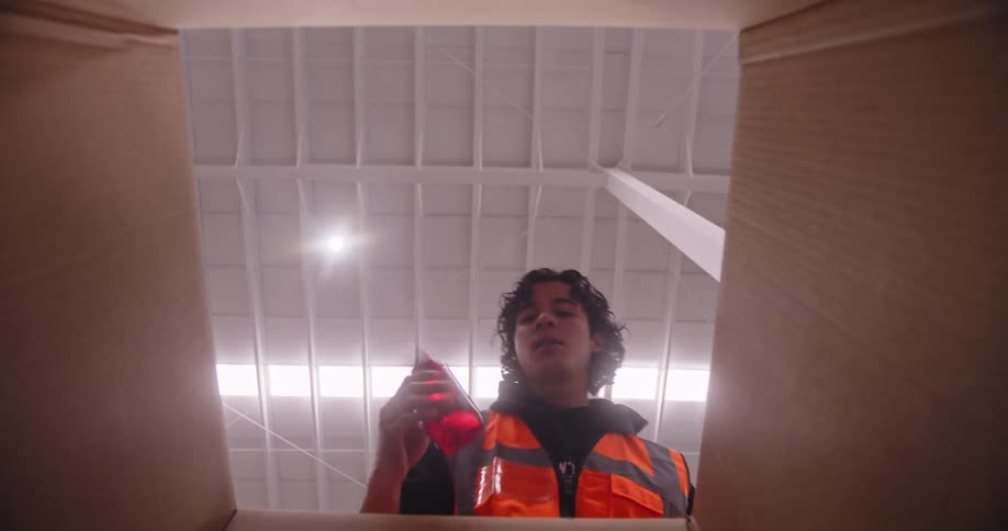 Low angle shot from inside a cardboard box of a warehouse employee carefully packing red bottles and closing the lid