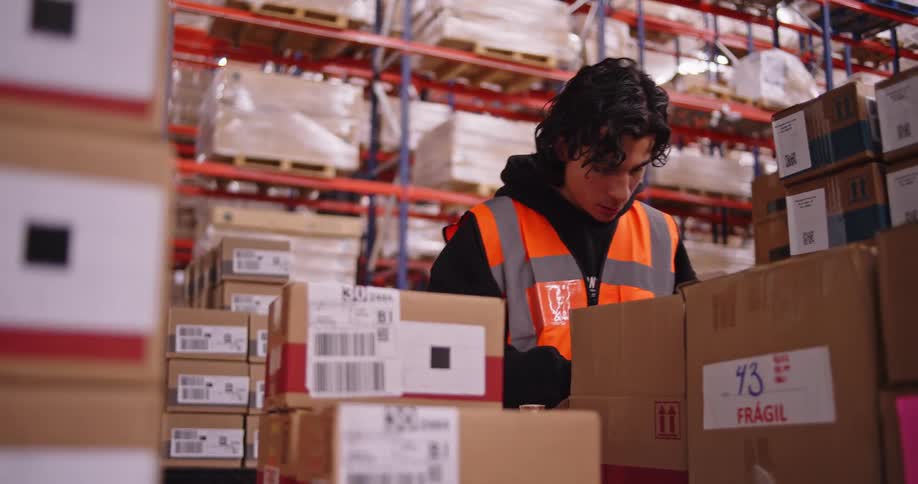 A young male warehouse employee carefully packs products into a cardboard box for shipping in a large distribution center