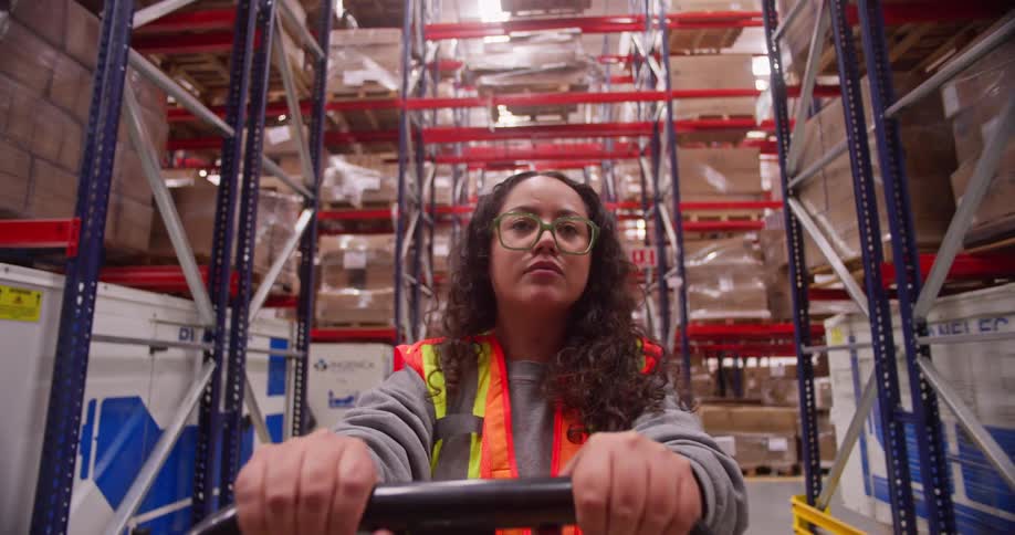 A point-of-view shot of a female warehouse worker operating a pallet jack down a long aisle filled with shelves of inventory