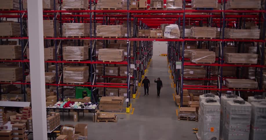 Two warehouse workers walk through a massive logistics center, inspecting inventory stacked high on industrial shelves