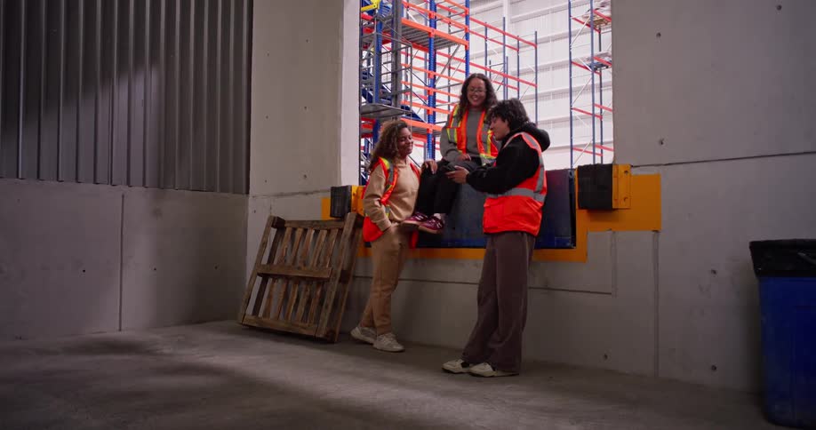 Three cheerful female warehouse workers in safety vests enjoy a friendly chat and laugh together during their break at the loading dock