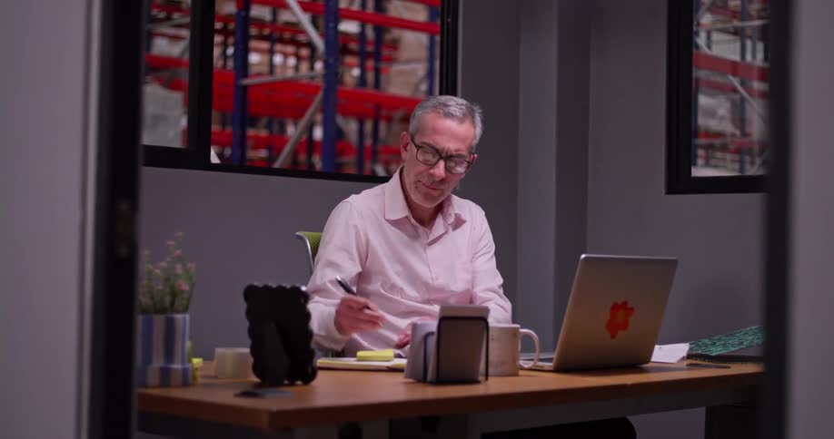 A focused businessman works diligently on his laptop and takes notes at his desk in an office with a view of a warehouse