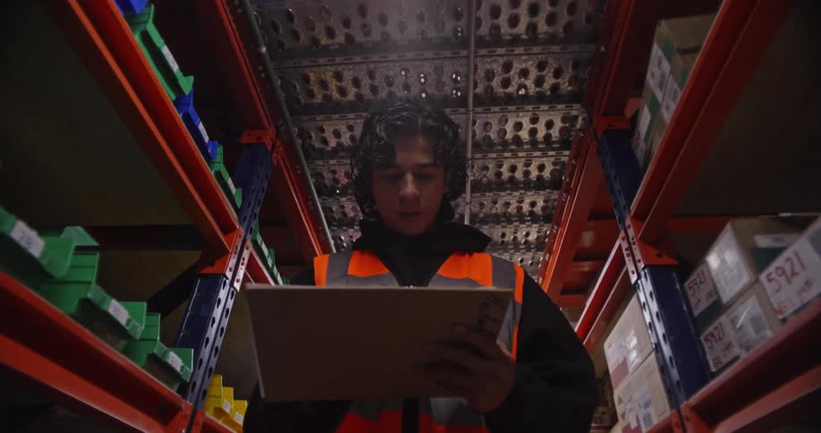 A young warehouse worker in a high-vis vest meticulously checks inventory, marking boxes and updating his clipboard while walking down a well-stocked aisle