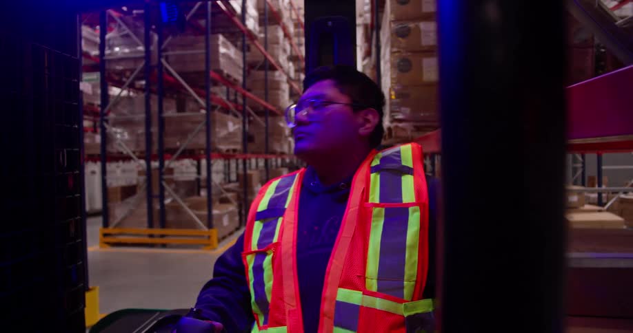 A focused forklift operator in a safety vest skillfully maneuvers the vehicle between tall shelves of inventory in a large warehouse