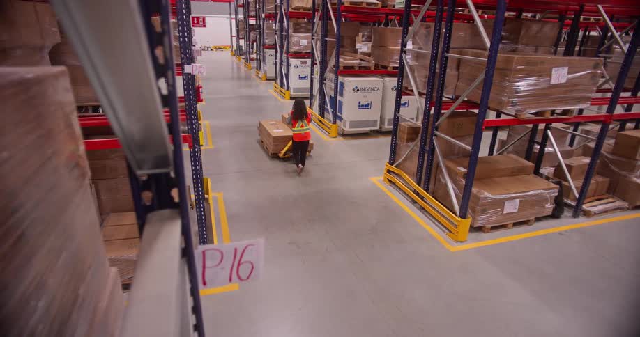 High-angle view of a female worker using a pallet jack to move boxes through the aisles of a large, modern warehouse