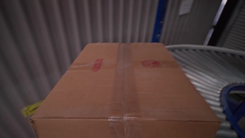 A point-of-view shot follows a cardboard box as it travels along an automated conveyor belt system inside a large industrial warehouse