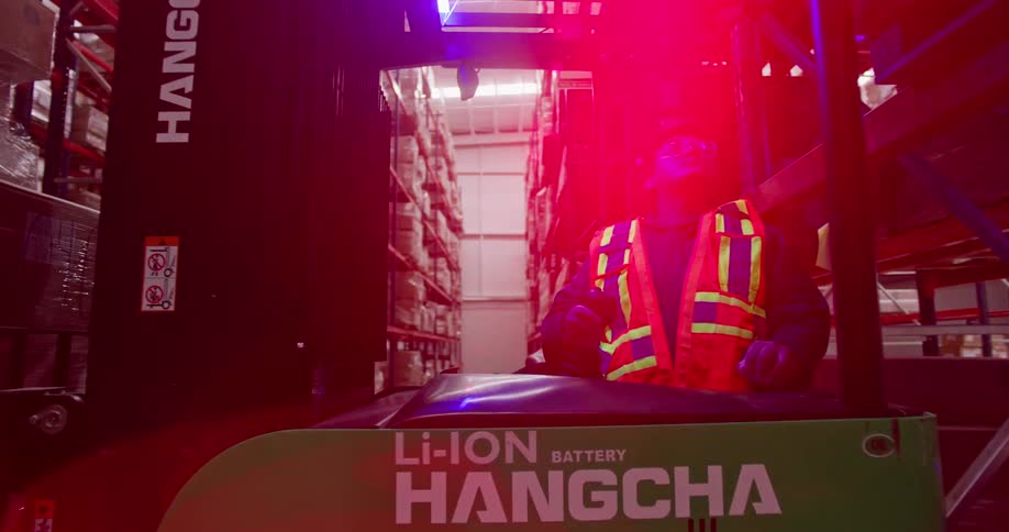 A forklift operator carefully maneuvers down a warehouse aisle, lifting and placing a large pallet of boxes onto a high industrial shelf
