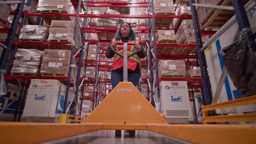 Point-of-view shot from a pallet jack as a female worker pulls it through a vast warehouse aisle