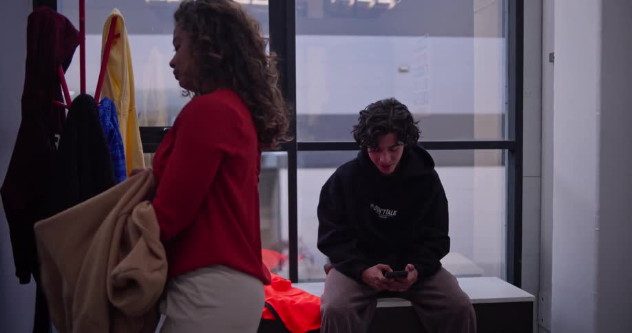 A woman joins her colleague in the break room, grabbing her hoodie from a locker while they engage in a friendly conversation