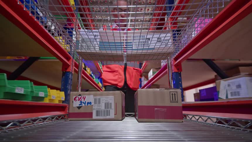 A unique low-angle POV shot from inside a warehouse cart as a worker pushes it down an aisle, scanning and organizing packages on shelves
