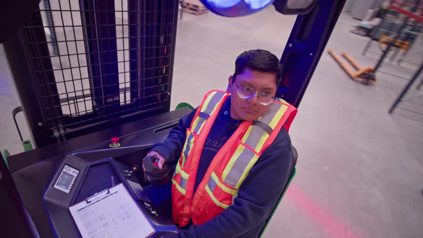 From a high-angle view, a serious warehouse worker operates a forklift, moving through the facility's aisles