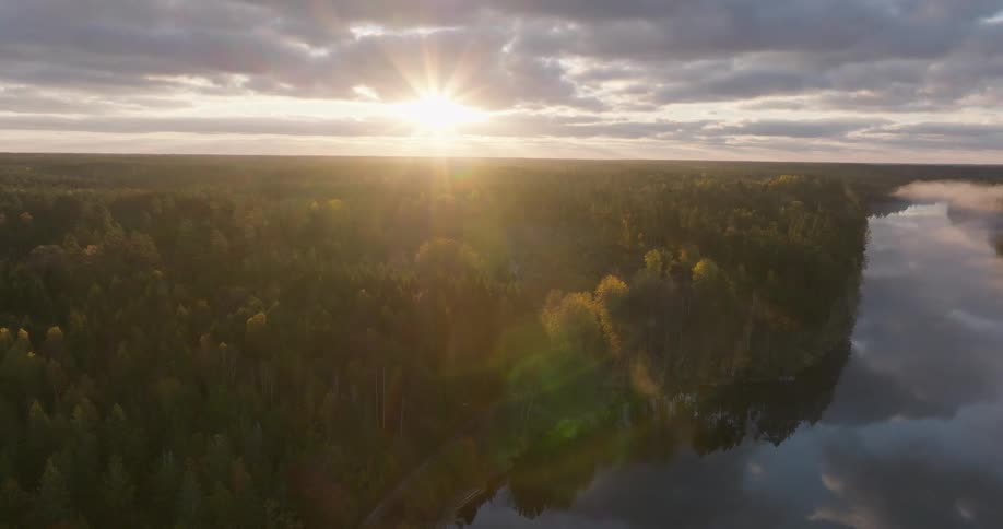 A stunning aerial view captures the sun rising over a misty autumn forest bordering a tranquil, reflective lake