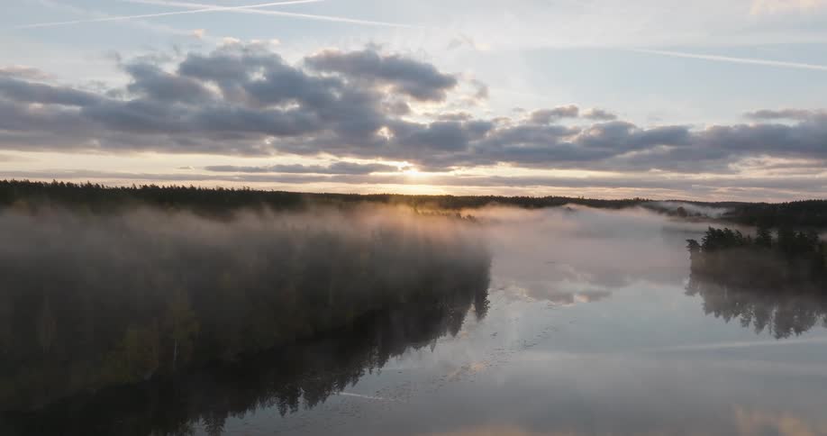 A breathtaking aerial view of a misty forest and calm lake at sunrise, with golden sunbeams piercing through the clouds and fog