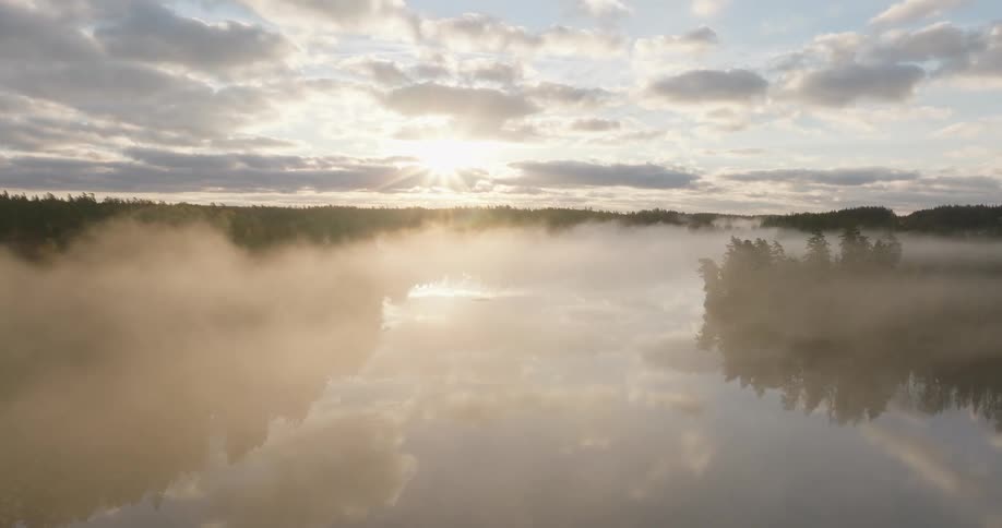 An ethereal aerial view reveals a tranquil lake shrouded in a thick layer of morning mist as the sun rises over a vast forest