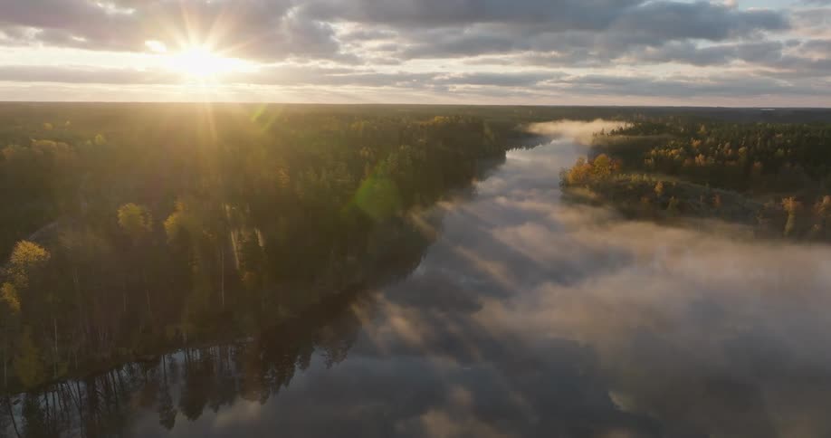 An aerial view shows a stunning sunrise over a tranquil river, with golden rays piercing through clouds and fog above a vast autumn forest