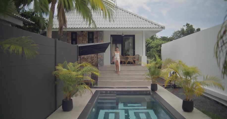 A slow-motion dolly zoom out reveals a beautiful woman in a white dress walking out of a luxurious tropical villa towards her private swimming pool