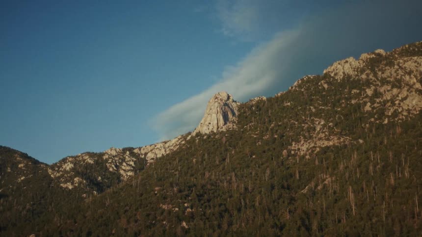 A majestic, tree-covered mountain with a prominent rock peak stands against a clear blue sky as clouds slowly drift by