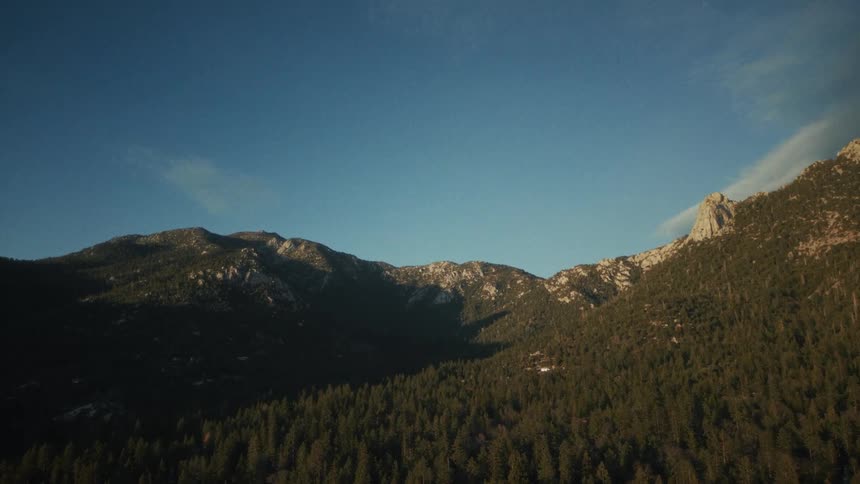 An aerial view tilts down from a clear blue sky to reveal a majestic, tree-covered mountain range during golden hour