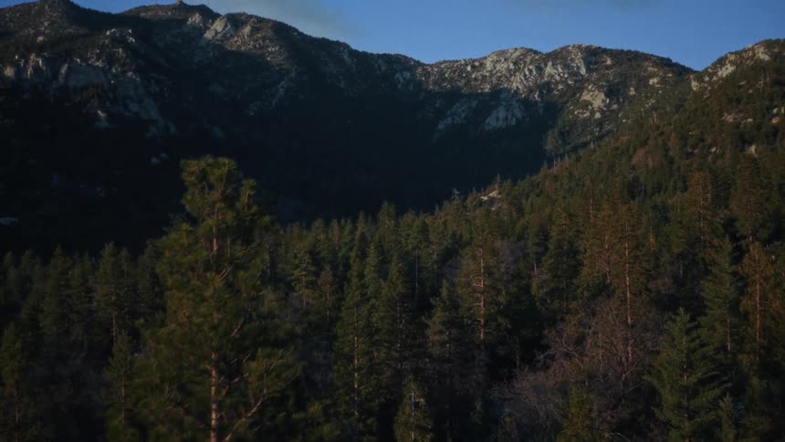 A slow aerial ascent reveals a vast, rocky mountain range towering over a dense evergreen forest under a clear blue sky
