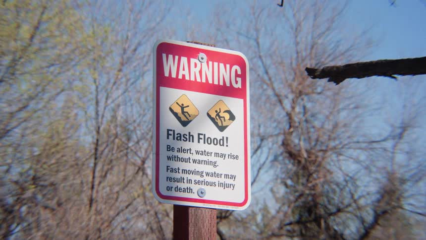 A slow zoom reveals a flash flood warning sign with a bullet hole posted in a dry, brushy landscape