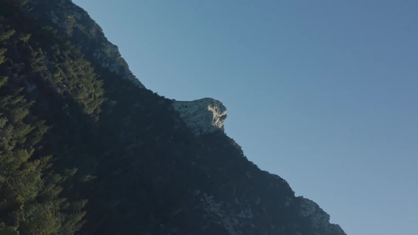 A vertical drone shot slowly reveals a sun-kissed rock peak on a majestic, forested mountain under a clear blue sky