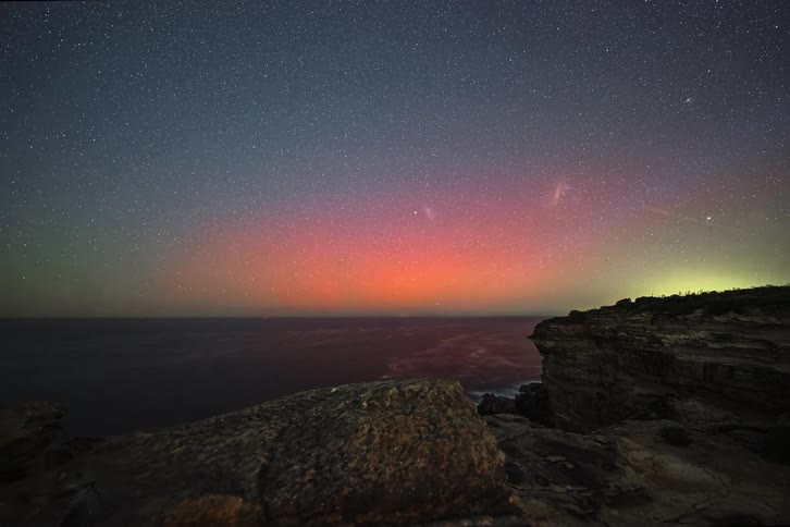 A stunning timelapse of the colorful Aurora Australis glowing on the horizon over the ocean, viewed from a rocky cliff under a star-filled night sky