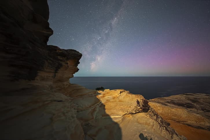 A stunning day-to-night timelapse from a rocky cliff reveals the vibrant Milky Way and the colorful Aurora Australis appearing over the ocean