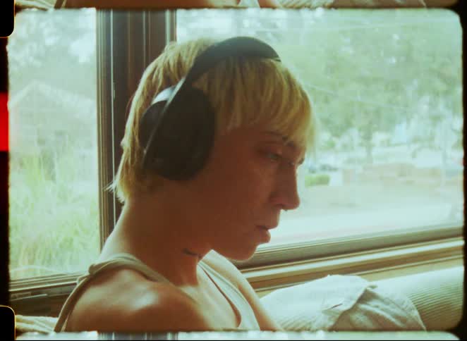 A thoughtful person with short blonde hair listens to music on headphones by a window in a vintage film-style clip